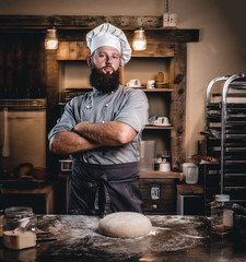 Professional baker in cook uniform posing with crossed arms near table with ready dough in the...