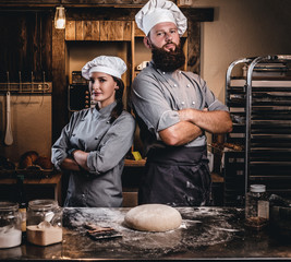 Chef with his assistant in cook uniform posing with crossed arms near table with ready dough in the bakery.