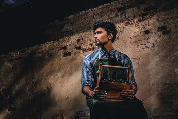 Tired brewer in apron sitting on a barrel and holds a wooden box with craft beer.