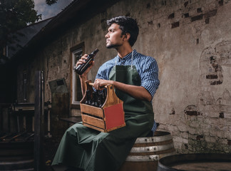 Brewmaster sitting on a wooden barrel and holds a glass of craft beer, relaxes after work.