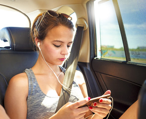 A cute teenage girl looking at the screen of her smartphone while sitting in a van wearing...