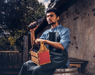 Brewmaster sitting on a wooden barrel and holds a glass of craft beer, relaxes after work.