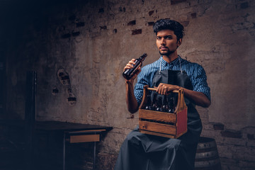 Brewmaster sitting on a wooden barrel and holds a glass of craft beer, relaxes after work.