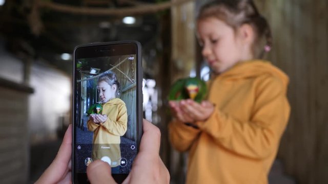 Mother takes smartphone pictures of little child girl daughter holding feeding a parrot bird