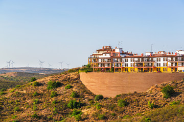Obraz premium Apartments built high on a hillside in Costa Esuri an urbanisation in Ayamonte, Alongside the River Guadiana in Andalusia, Spain. Turbines in a wind farm can be seen on the hillside