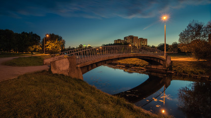 a pedestrian concrete bridge with metal handrails over a narrow river in the autumn evening city park is illuminated by the light of street electric lamps