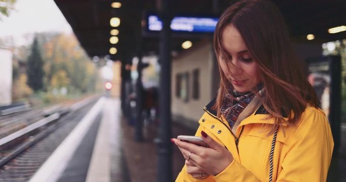 Young Woman Using Smartphone at the Train Station. SLOW MOTION 4K. Girl using cell phone, waiting for city train. Social network, planning, communicating.
