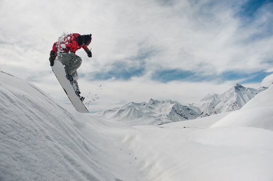 Jumping snowboarder keeping his hand on the snowboard on the background of mountains and sky