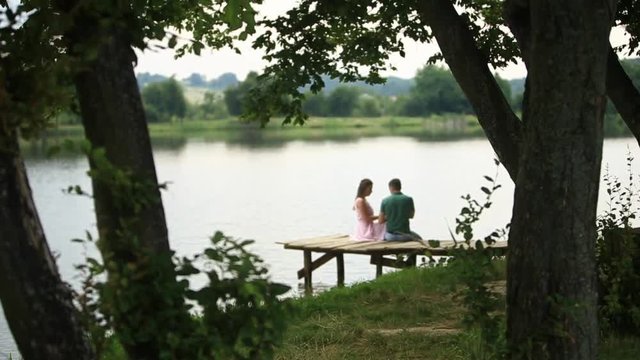 Beautiful youn couple sit on the pier near the lake. Romantic atmosphere. Back biew