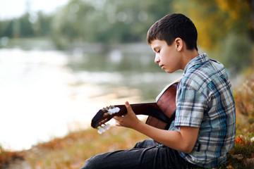the boy plays an acoustic guitar, sits on the Bank of the river, autumn forest at sunset, beautiful nature and the reflection of trees in the water