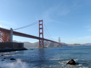 Naklejka premium Wave breaks on shore with Golden Gate Bridge, Marin County and Fort Point in distance