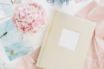 Home office desk with family wedding photo album, pastel colorful hydrangea flower bouquet, peachy blanket, decoration on white background. Flat lay, top view.