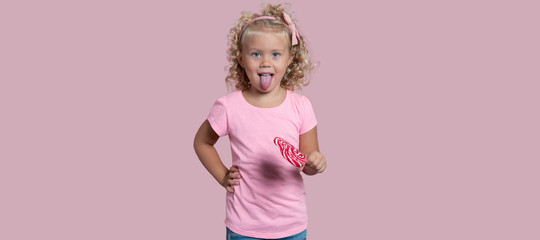 Fototapeta premium Small blond girl holds lollipop and shows a tong isolated over pink background