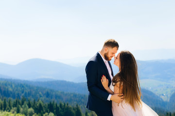 the groom in a suit hugs a bride with long hair and a white dress against the background of the mountains