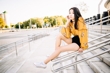 Asian beautiful woman with black hair, yellow shirt, black shorts, outdoor resting, looking at camera. Portrait young girl in an urban environment.