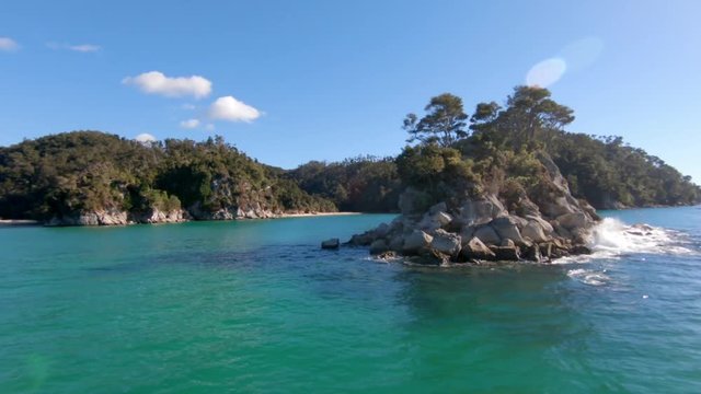 View on a small island rocky island with a few trees on it in the ocean. In the background are lush green hills of the coastline