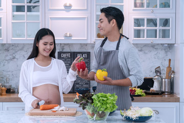 Attractive cheerful young husband looking his pregnant wife while cooking vegetables food together in the kitchen. Concept healthy food, happy family.