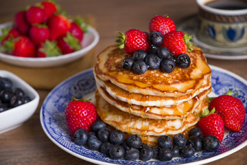 Stack of pancakes with blueberries, strawberries, and maple syrup on a blue and white plate with bowls of strawberries and blueberries and a cup of coffee in the background. 
