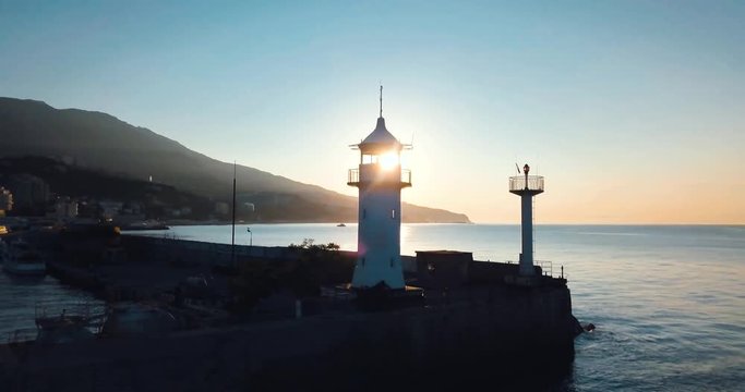Lighthouse for ships and sunrise over the sea. Epic on the edge of the mountain valley. Sun flare. 4k drone flight. Aerial establisher. City by the ocean. Concept of marine travel. Film vintage colors