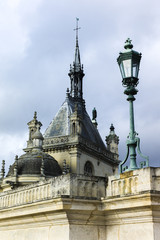 Obraz premium Lamp post and Palace of Chantilly in golden afternoon light. Rooftops and sky view