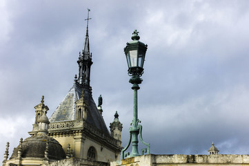 Fototapeta na wymiar Lamp post and Palace of Chantilly in golden afternoon light. Rooftops and sky view