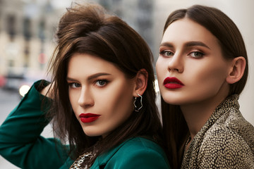 Outdoor close up fashion portrait of two young beautiful women with red lips makeup, wearing trendy clothes and accessories,  posing in street of european city