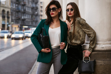 Outdoor fashion portrait of two young beautiful women wearing trendy sunglasses, stylish clothes, holding black leather bags, posing in street of european city. Copy, empty space for text - obrazy, fototapety, plakaty