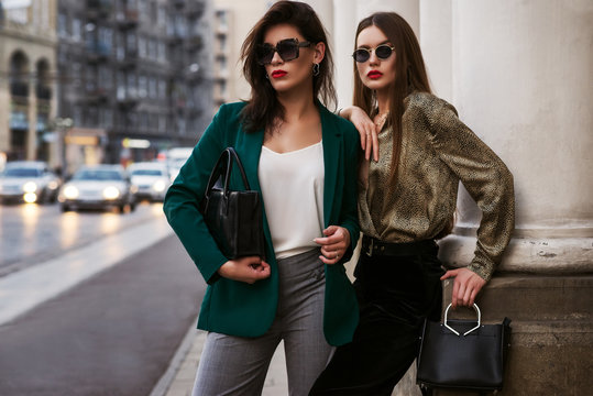 Outdoor fashion portrait of two young beautiful women wearing trendy sunglasses, stylish clothes, holding black leather bags, posing in street of european city. Copy, empty space for text
