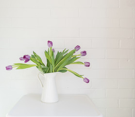 Purple tulips in white jug on table against painted brick wall with copy space