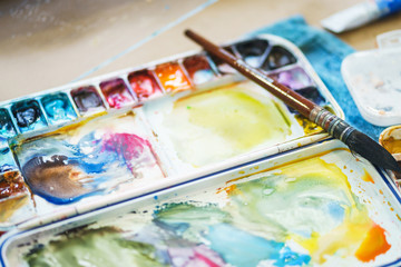 Palette with watercolors in different colors. Various brushes and pencils for drawing prepared by the artist for work.