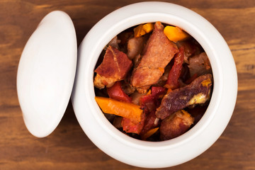 Ceramic white pot with lid in which cooked meat stew with vegetables, wooden background.