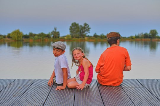 Children sitting on pier. Three children of different age - teenager boy, elementary age boy and preschool girl sitting on a wooden pier. Summer and childhood concept. Golden hour during evening