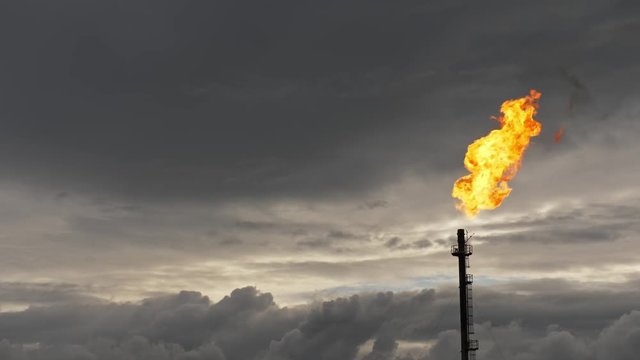 A tall GAS TORCH burns with a vivid flame against a moody dusk sky at an oil refinery. Industrial landscape under dense clouds.