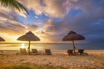 Die Sonne geht über dem Meer bzw. dem Ozean vor Mauritius unter. Sonnenliegen und geflochtene Schirme zeigen des Urlaubsidyll.