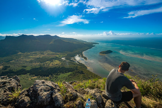 Der Berg ist erklommen, auf dem Gipfel rastender Wanderer blickt auf das t&uuml;rkise Meer und die gr&uuml;ne Insel namens Mauritius bei herrlichem Sonnenschein und gro&szlig;er Hitze