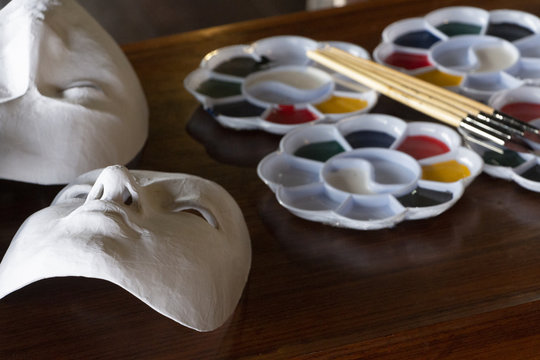 White paper Venetian-style masks lying next to watercolor dish filled with bright colors with brushes placed over them before getting painted on