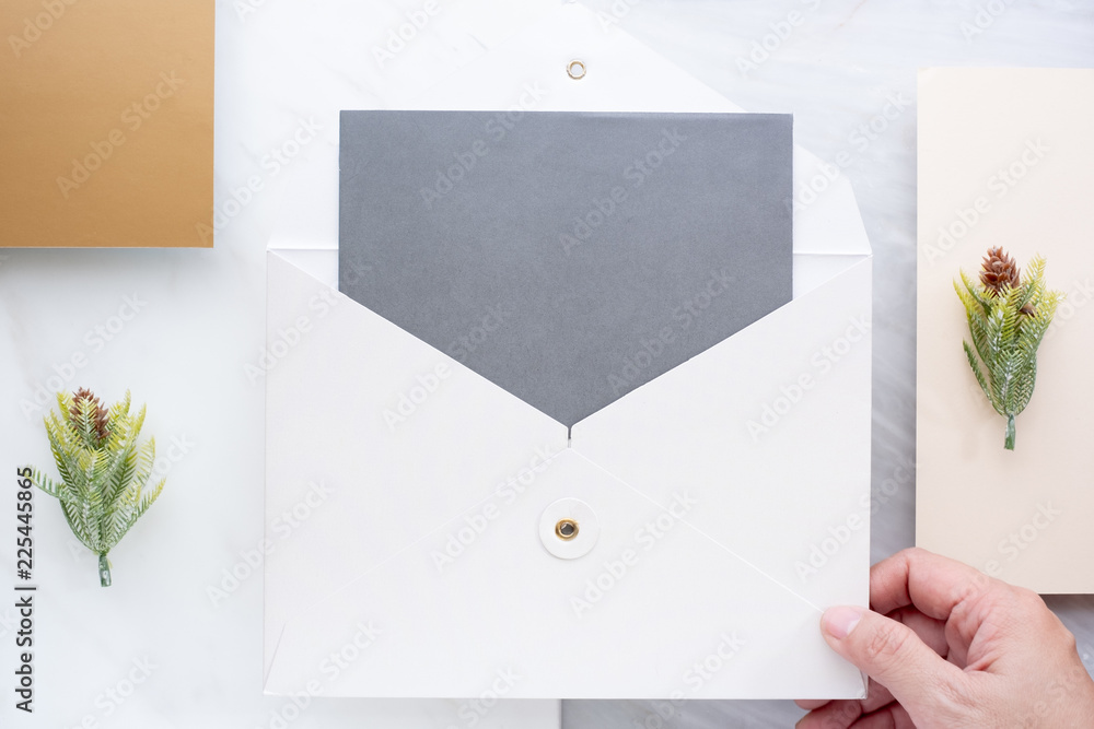 Poster Top view of hand holding blank grey card in white envelop with pine leaf on two layer step of marble table top.Mock up template for display of design.modern business items.