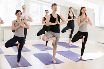 Fototapeta premium Group of young sporty people practicing yoga lesson, doing Vrksasana exercise, Tree pose, working out, indoor, students training in club, studio full length. Well-being concept