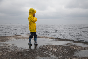  A boy in a yellow raincoat is standing in a puddle on the sea in cloudy weather.