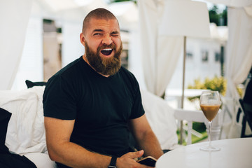 Young bearded tired man yawning, drinking whipped coffee with cream (Italian caffè shakerato) sitting in a beach lounge during vacation in Italy - laziness, relax, healthy mediterranean food