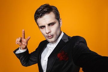 Photo of scary zombie man on halloween wearing classical suit and creepy makeup taking selfie,...