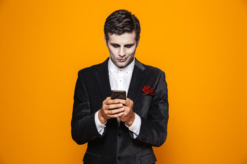 Photo of european dead man on halloween wearing classical suit and creepy makeup holding mobile phone, isolated over yellow background