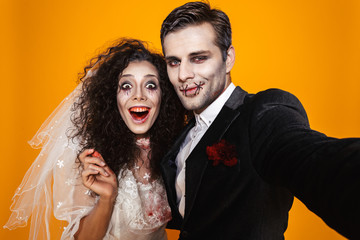 Photo of amusing zombie couple bridegroom and bride wearing wedding outfit and halloween makeup laughing while taking selfie, isolated over yellow background