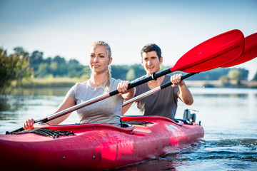 Happy young couple holding paddle kayaking together on lake - obrazy, fototapety, plakaty