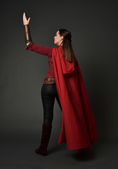 full length portrait of brunette girl wearing red medieval costume and cloak. standing pose  with back to the camera on grey studio background.