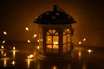 Fototapeta na wymiar Glowing lantern in the shape of a house with Windows lit yellow. Festive interior decoration for the New year. Lights garlands and candles inside the lamp, glowing stars on the roof