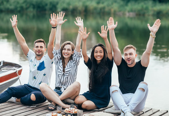 Happy friends raised hands relaxing on picnic outdoors on river
