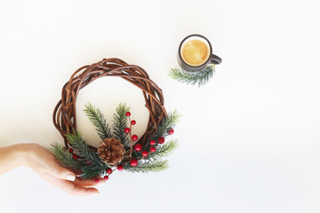 Christmas wreath with green branches of a Christmas tree and red berries in a female hand next to a cup of coffee. Festive minimalism. Top view, flat lay