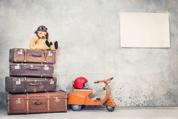 Retro Teddy Bear toy in aviator's hat, aged travel valises, children's pedal scooter, helmet, paper poster blank on concrete wall background. Vintage style filtered photo