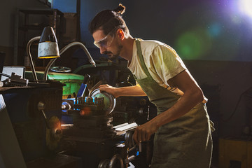 concentrated male manufacture worker in protective apron and goggles using machine tool at factory